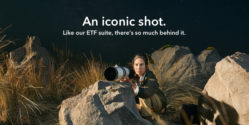 A photograph of a woman crouched behind a boulder with a large, professional camera, waiting for the perfect shot. The text says: An iconic short. Like our ETF suite, there's so much behind it.