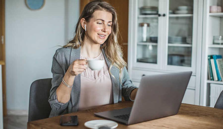 A woman sitting at a desk. She is dressed in a blazer and holding a cup of cup of coffee. She is smiling while speaking with someone on her laptop. 