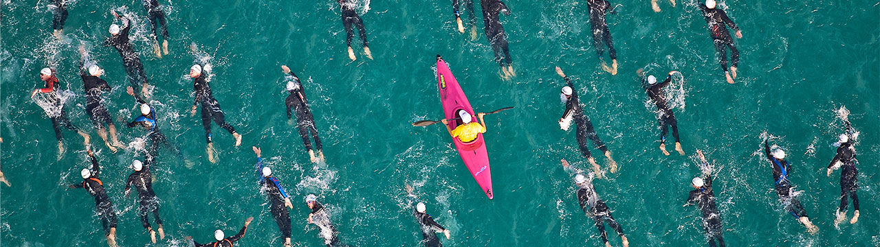A group of swimmers swimming in the ocean dressed in black swimsuits and white caps. In the middle of this large group is a pink kayak. The person inside the kayak is rowing and dressed in a yellow shirt and a white cap.