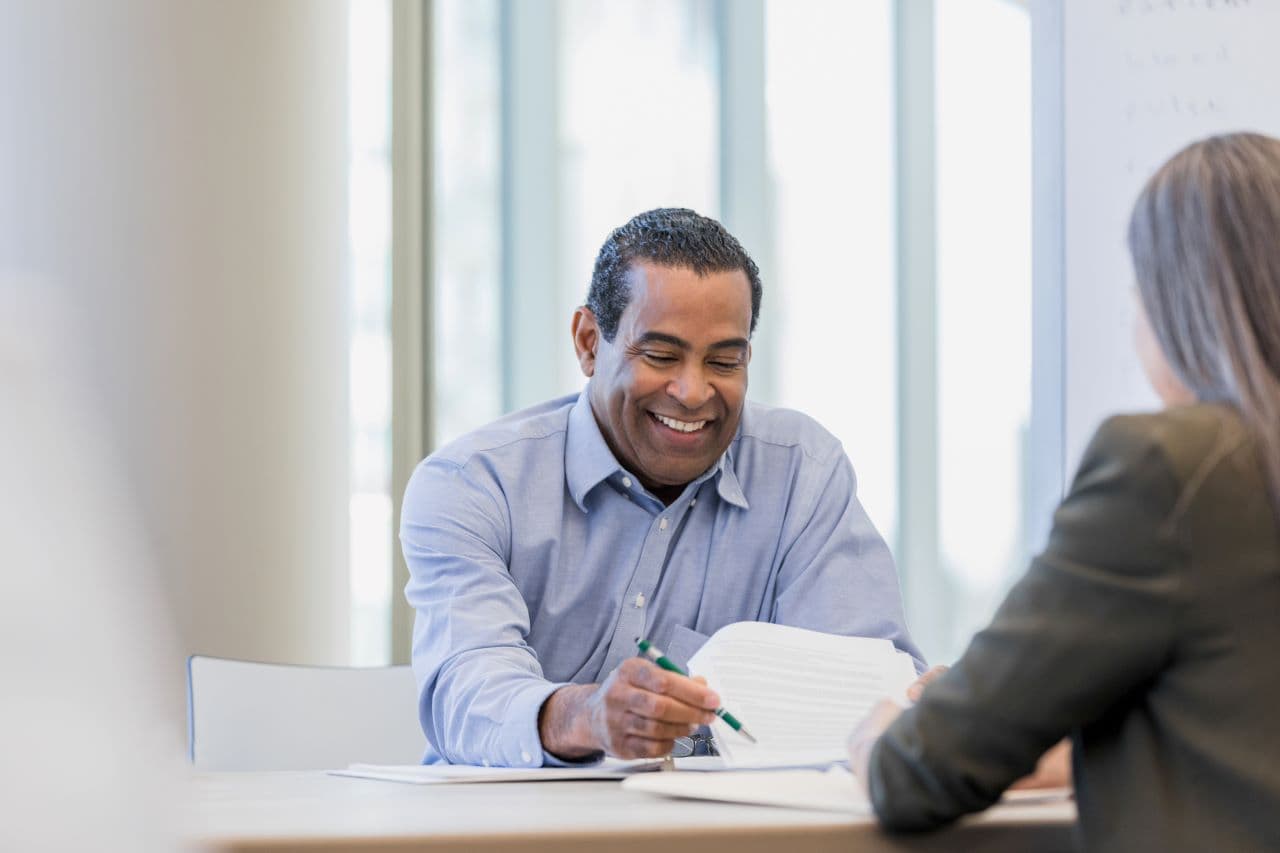 Man smiles as he looks at paperwork as a woman sitting opposite leans forward. 