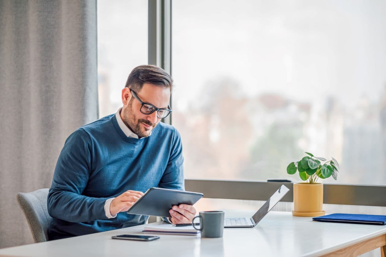 Man with brown hair sitting at a desk wearing a blue shirt and eye glasses looking at a tablet.