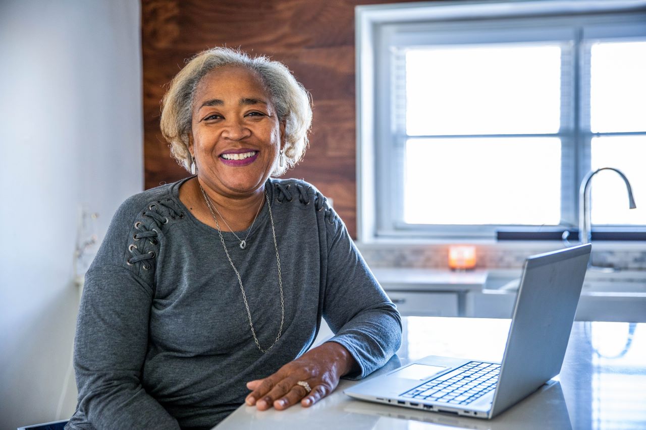 A woman with grey hair and a grey shirt smiles toward the camera as she leans on her desk. Her laptop is open in front of her.